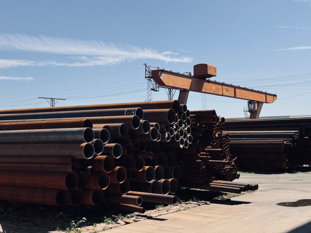 Stacked steel pipes and an overhead crane in an industrial yard under a clear blue sky.