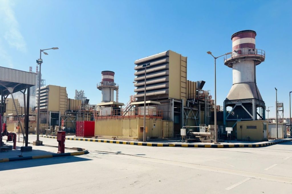 A modern power plant with turbines and chimneys under a clear blue sky in Saudi Arabia.
